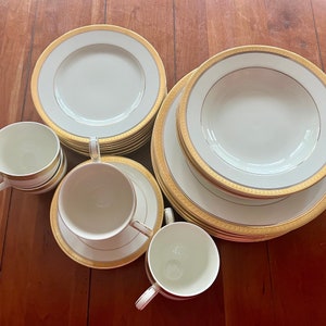 May include: A set of white porcelain dinnerware with gold trim. The set includes a large dinner plate, a smaller salad plate, a soup bowl, and two teacups and saucers.