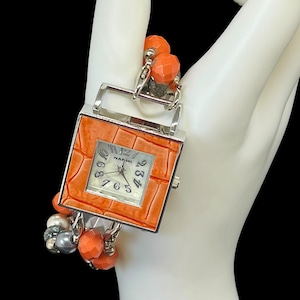 Puede incluir: Un reloj cuadrado naranja con marco plateado y esfera texturizada. La esfera del reloj tiene manecillas y números plateados. El reloj está sujeto a una pulsera de cuentas naranjas, grises y plateadas. Es un accesorio de moda.
