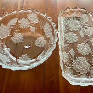 Puede incluir: Dos platos de servir de vidrio transparente con un diseño floral. El plato más grande es redondo y tiene un borde festoneado. El plato más pequeño es rectangular. Ambos platos tienen un patrón floral en relieve.
