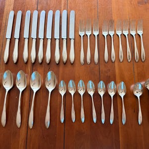 May include: A large set of polished stainless steel flatware including nine dinner knives, eight dinner forks, five large spoons, eight smaller spoons, one soup ladle, and one butter knife, all featuring a smooth, tapered handle design.