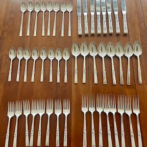 Puede incluir: Un conjunto de cubiertos de plata antiguos, que incluye cucharas, tenedores y cuchillos, dispuestos sobre una superficie de madera. La cubertería tiene un acabado plateado con detalles decorativos en los mangos. La disposición sugiere una mesa formal.