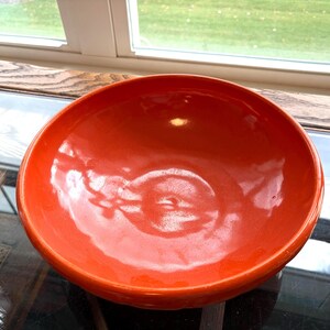 May include: A round, deep ceramic bowl with a smooth, glossy finish in a vibrant orange color. The bowl features a wide, curved rim and a solid, uniform orange interior and exterior, reflecting light on its polished surface.