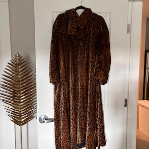 May include: A long-sleeved, calf-length coat featuring a brown and black leopard print pattern. The garment is crafted from a velvet-like fabric and includes a wide, folded collar, cuffed sleeves, and a button-down front closure.