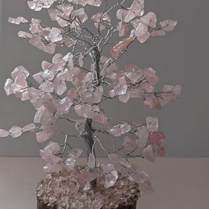 May include: A decorative tree sculpture featuring a silver wire trunk and branches, embellished with light pink rose quartz chips. The tree is set in a small, round, wooden base, creating a natural and elegant aesthetic.