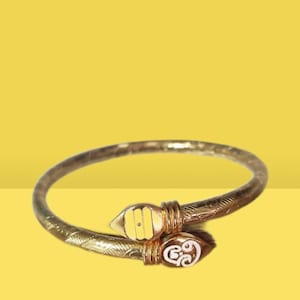 May include: A gold-colored bracelet with an open design. The bracelet features decorative detailing along the band and at the ends. The ends have white and yellow accents. The bracelet is set against a yellow background.