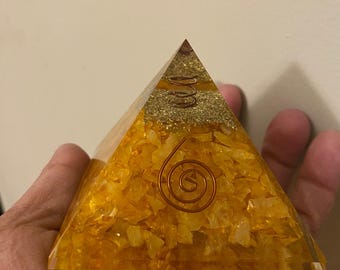 May include: A transparent, yellow resin pyramid with embedded yellow crystals and a copper coil. The pyramid has a layered design with a gold glitter layer. The pyramid is held in a hand.