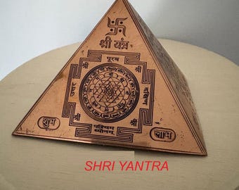 Könnte beinhalten: Eine Kupferpyramide mit einem eingravierten Shri-Yantra-Design. Die Pyramide steht auf einer Holzoberfläche. Der Text auf der Pyramide lautet "Shri Yantra" und enthält Sanskrit-Zeichen.