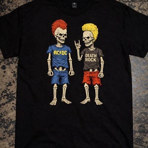 May include: Black t-shirt featuring a cartoon illustration of two skeleton figures. One skeleton wears a blue shirt and shorts with "AC/DC" text. The other skeleton wears a black shirt and red shorts with "DEATH ROCK" text.