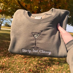 May include: Olive green sweatshirt with an embroidered martini glass design. The words "Dirty, but Classy" are embroidered below the martini glass. The sweatshirt is held up in front of a background of fall foliage.