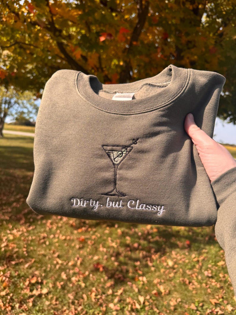 May include: Olive green sweatshirt with an embroidered martini glass design. The words "Dirty, but Classy" are embroidered below the martini glass. The sweatshirt is held up in front of a background of fall foliage.