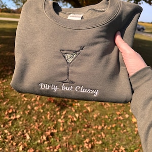 May include: Olive green sweatshirt featuring an embroidered martini glass design. The words "Dirty, but Classy" are embroidered below the martini glass. The sweatshirt is held up in front of a background of grass and trees.