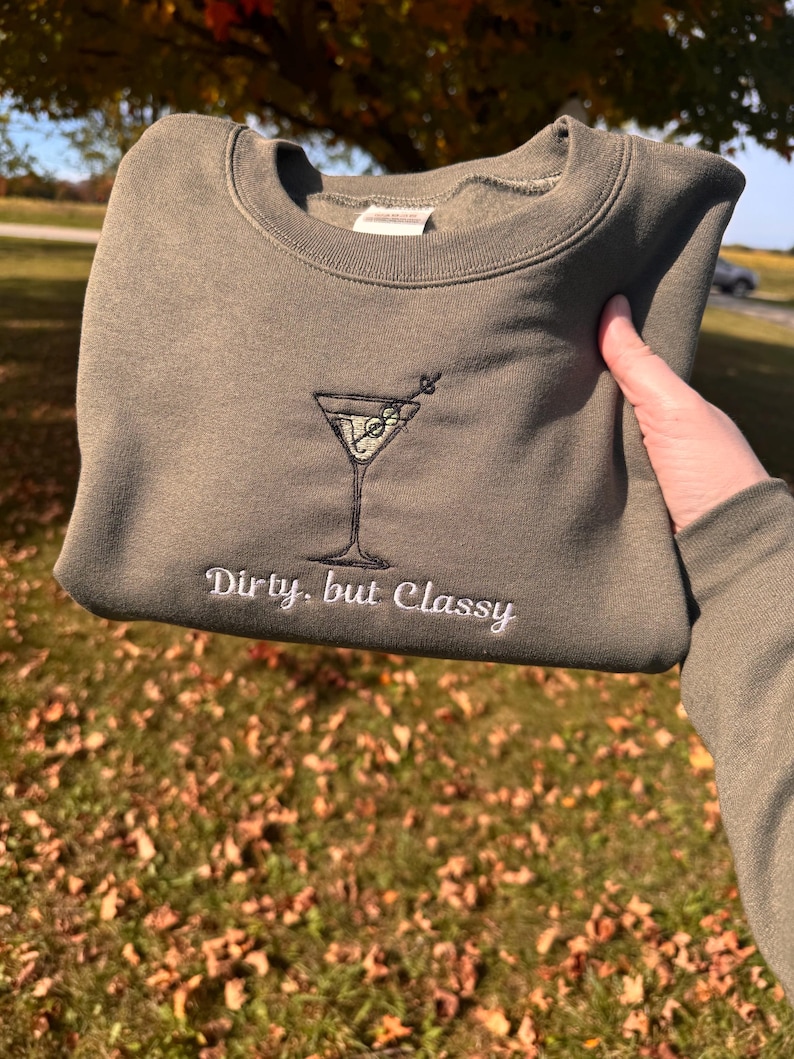 May include: Olive green sweatshirt featuring an embroidered martini glass design. The words "Dirty, but Classy" are embroidered below the martini glass. The sweatshirt is held up in front of a background of grass and trees.