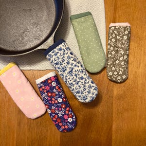 May include: A collection of colorful cast iron skillet handle covers. The covers feature various floral patterns, polka dots, and solid colors, including pink, blue, green, and brown. The covers are designed to protect hands from heat.