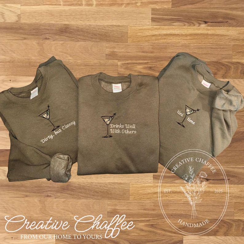 May include: Three olive green sweatshirts with embroidered martini glass designs. One says "Dirty, but Classy," another "Drinks Well With Others," and the third "tini time." The sweatshirts are from Creative Chaffee.