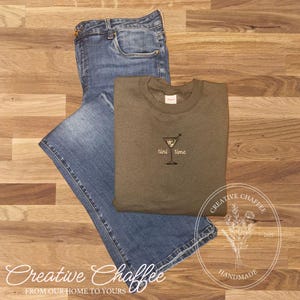 May include: An olive green sweatshirt with the embroidered text "tini time" next to a martini glass, paired with blue denim jeans. The items are arranged on a wooden surface. The image also includes the text "Creative Chaffee" and "Handmade".