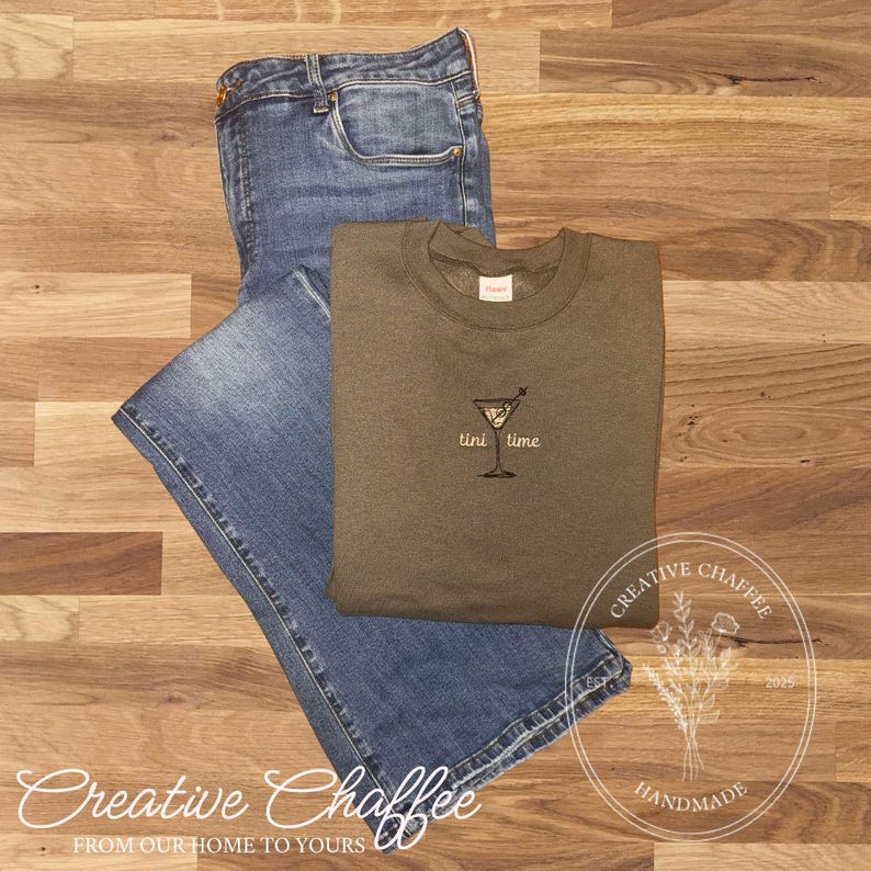 May include: An olive green sweatshirt with the embroidered text "tini time" next to a martini glass, paired with blue denim jeans. The items are arranged on a wooden surface. The image also includes the text "Creative Chaffee" and "Handmade".