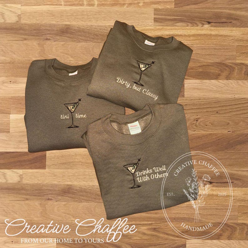 May include: Three olive-green sweatshirts with embroidered martini glass designs. One says "tini time," another "Dirty, but Classy," and the third "Drinks Well With Others." The sweatshirts are on a wooden surface.