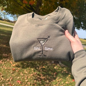 May include: Olive green sweatshirt with an embroidered martini glass design and the words "tini time" in white. The sweatshirt is folded and held up against a background of fall foliage.