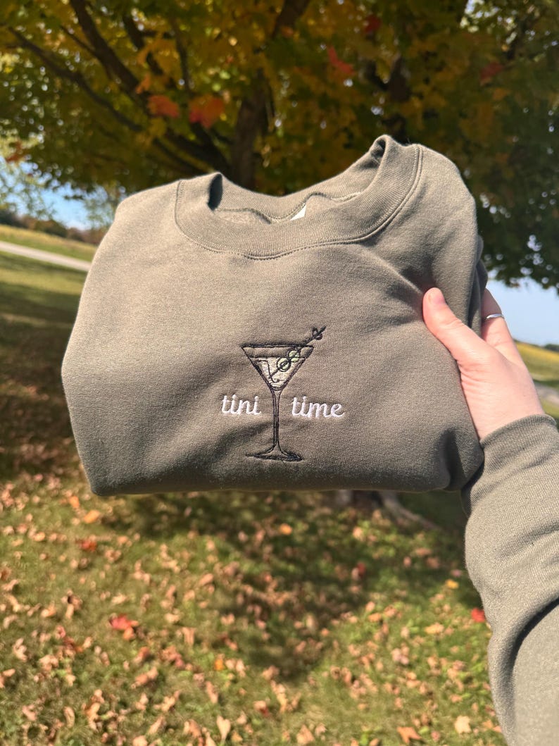 May include: Olive green sweatshirt with an embroidered martini glass design and the words "tini time" in white. The sweatshirt is folded and held up against a background of fall foliage.