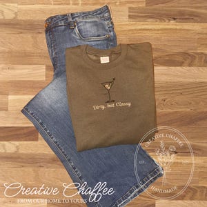 May include: An olive green sweatshirt with an embroidered martini glass design and the text "Dirty, but Classy." The sweatshirt is paired with blue jeans on a wooden surface. The image also includes the text "Creative Chaffee" and "FROM OUR HOME TO YOURS."