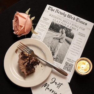 以下が含まれることがあります： 「The Newly Wed Times」というタイトルの新聞と「Wedding Bells Ring for Clayton & Emily」という見出しがある結婚式のシーン。チョコレートケーキとフォークの入ったお皿、バラ、キャンドル、そして「Just Married」と書かれたナプキンも写っています。