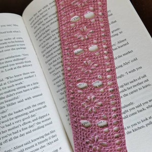 Op de afbeelding: Een handgemaakte, open geweven boekenlegger in een stoffige roze kleur, met een gefranjerde rand. De boekenlegger ligt op de pagina's van een boek en voegt een vleugje elegantie toe aan het lezen.