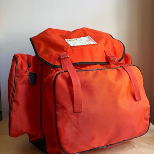 May include: A bright orange backpack featuring a large main compartment with a flap closure, two side pockets, and black piping trim. Two vertical orange straps secure the front flap, which displays a small rectangular white label with text.