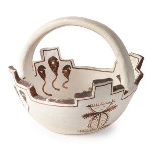 May include: A white ceramic basket with a brown handle and intricate brown designs. The basket has a rounded shape and a wide opening. The designs include stylized figures and geometric patterns.