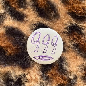 May include: A white button with a purple handwritten number "999" on it. The button is on a brown and black fuzzy surface.