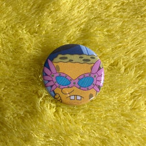 May include: A round button featuring a cartoon image of Spongebob Squarepants wearing pink and blue glasses. The button has a yellow background and is set against a yellow, fuzzy surface.