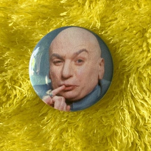 May include: A round button with a picture of a bald man with a serious expression. He is wearing a blue shirt and has his finger to his lips.