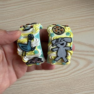 May include: Two decorative, colorful, cartoon-themed magnets. One features a blue bird and a mouse, the other an elephant and a cookie. Both have yellow and white frosting-like designs with blue and green accents.