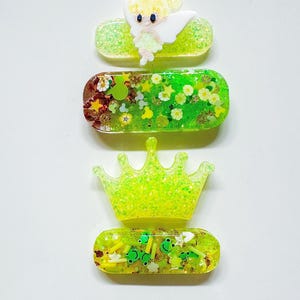 May include: Four green and gold glitter hair clips. One clip features a white fairy with yellow hair. Another clip has a green crown. The other two clips have green and gold glitter with various green and gold embellishments.