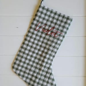 May include: A Christmas stocking featuring a green and white gingham check pattern. The name Violet is embroidered in red cursive thread across the upper section of the fabric. A fabric loop is attached to the top corner for hanging purposes.