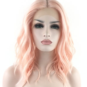 May include: A light pink wavy synthetic wig with a lace front. The wig is styled in a shoulder-length bob with loose waves.