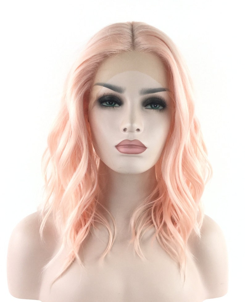 May include: A light pink wavy synthetic wig with a lace front. The wig is styled in a shoulder-length bob with loose waves.