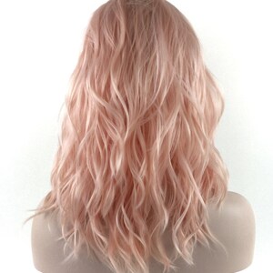 May include: A light pink wavy synthetic wig with a natural-looking parting. The wig is styled in a medium-length, layered cut.