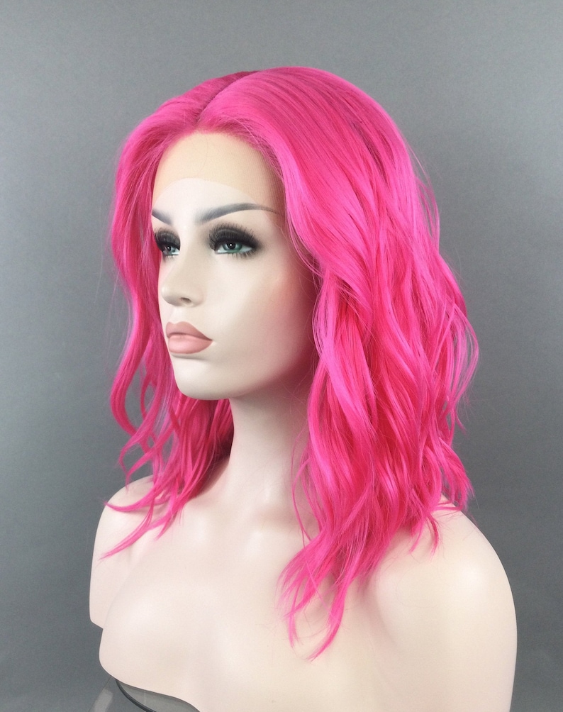 May include: A bright pink, wavy synthetic wig with a lace front. The wig is styled in a shoulder-length bob.