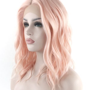 May include: A light pink wavy synthetic wig with a lace front. The wig is styled in a shoulder-length bob with loose waves.