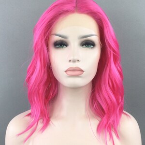 May include: A bright pink, wavy synthetic wig with a lace front. The wig is styled in a shoulder-length bob.
