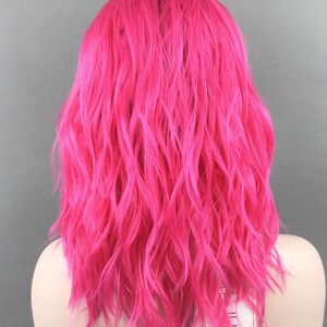May include: A bright pink wavy synthetic wig with a lace front. The wig is on a mannequin head and has a label that says "Illusion Lace Front".