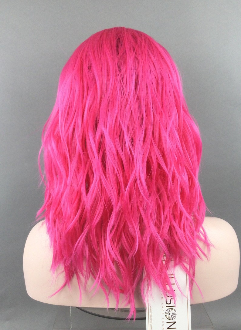 May include: A bright pink wavy synthetic wig with a lace front. The wig is on a mannequin head and has a label that says "Illusion Lace Front".