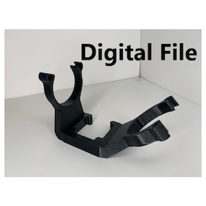 Peut inclure: Un support d'appareil numérique imprimé en 3D en plastique noir. Le support a une ouverture circulaire en haut et une base plate.