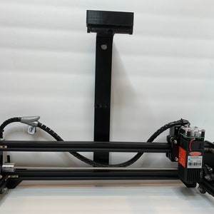 Puede incluir: Un grabador láser negro con un marco de metal negro y un módulo láser rojo. El grabador está montado en un soporte de metal negro fijado a una pared blanca.