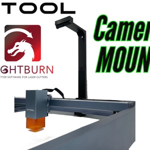 May include: A gray laser cutter with a camera mount attached to the top. The camera mount is black and has a rectangular shape. The laser cutter has a red and white logo that says "Lightburn" and a green and white logo that says "XTOOL". The text "Camera Mount" is written in green and black letters on a white background.