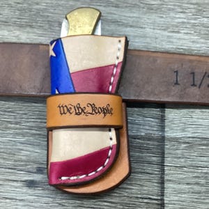 May include: A tan leather knife sheath featuring a red, white, and blue flag design with white stitching. A horizontal leather strap across the center is embossed with the text We The People. The sheath is positioned on a 1 1/2 inch brown belt.