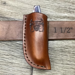 May include: A tan leather sheath with a stamped bull skull design, featuring visible stitching along the edge. The sheath is attached to a brown leather belt clearly marked with the measurement 1 1/2 inches, holding a small silver metal tool.