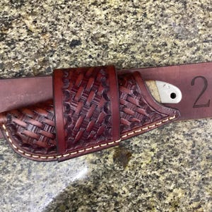 May include: A small knife with a light-colored handle is secured in a dark brown, intricately woven leather sheath. The sheath is attached to a matching brown leather belt, with the number "2" in black ink. The knife and sheath are approximately 2 inches in length.