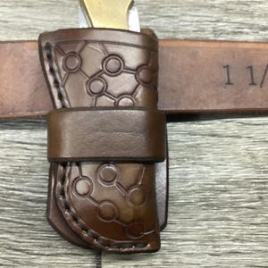 May include: A dark brown leather knife sheath featuring a geometric circle and line pattern, stitched edges, and a horizontal belt loop. The sheath is positioned on a brown leather belt clearly marked with the measurement 1 1/2 inches.