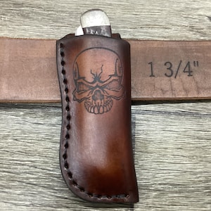 May include: A dark brown leather knife sheath featuring a stamped skull design on the front. The sheath is positioned over a brown leather belt with the text 1 3/4" stamped into the material, indicating the width of the belt strap.